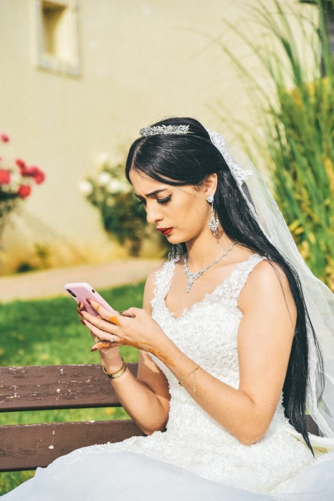 Woman in a wedding dress sitting on a bench looking at her mobile phone.